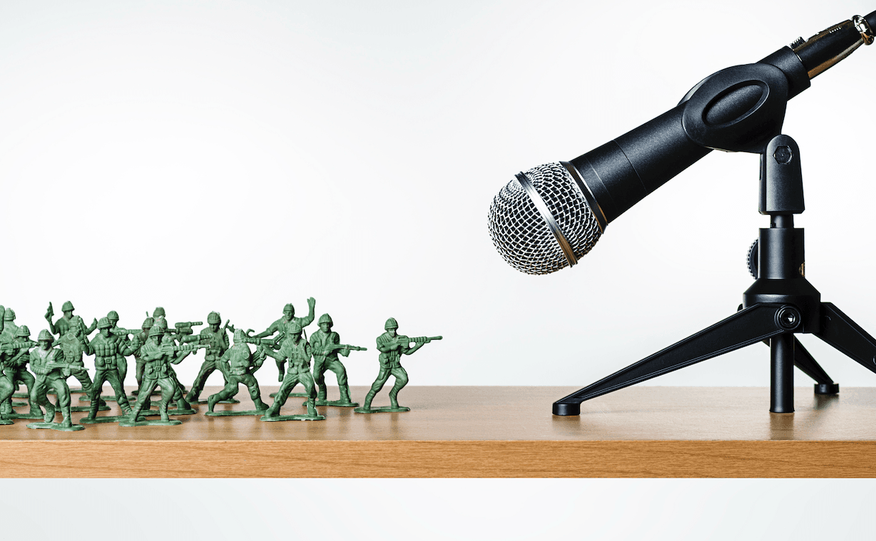 On a wood desktop, a cluster of green toy soldiers face a podcast microphone.