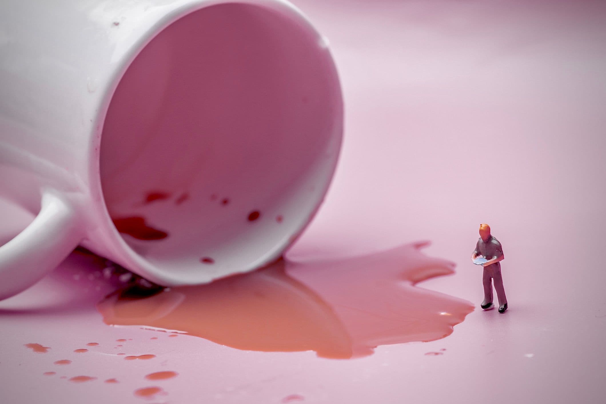 An image of a miniature human figure assessing spilled coffee from an overturned mug.