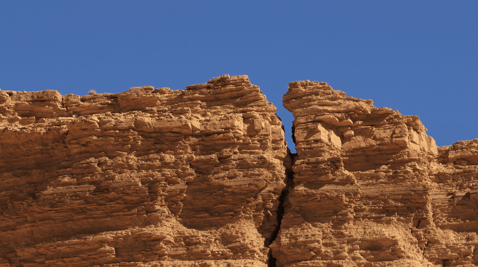 A photo of a big vertical crack in a rock cliff underneath a bright blue sky.