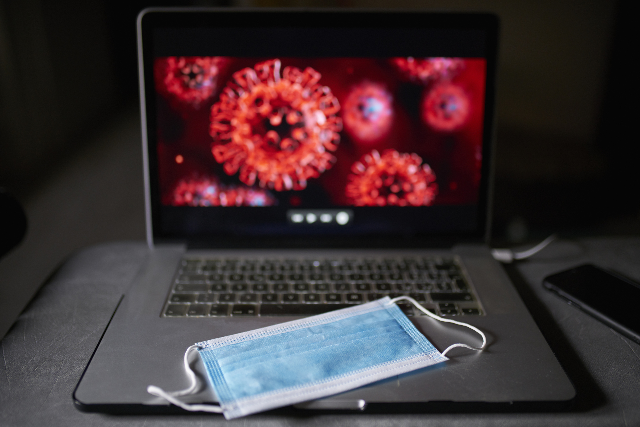 In a dark space, a blue face mask sits on a laptop keyboard, with microscopic images of the spiky red COVID virus glaring from the screen.