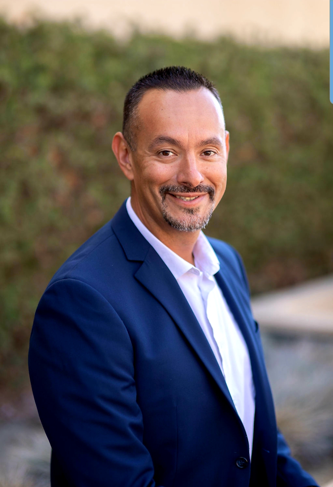 Photo of a man, 40-ish, with short black hair, tawny complexion, mustache and stubbled chin, displaying a warm smile.