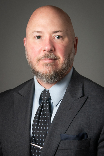 Photo of a bald White man in his 30s with a mustache and beard and wearing a gray suit.