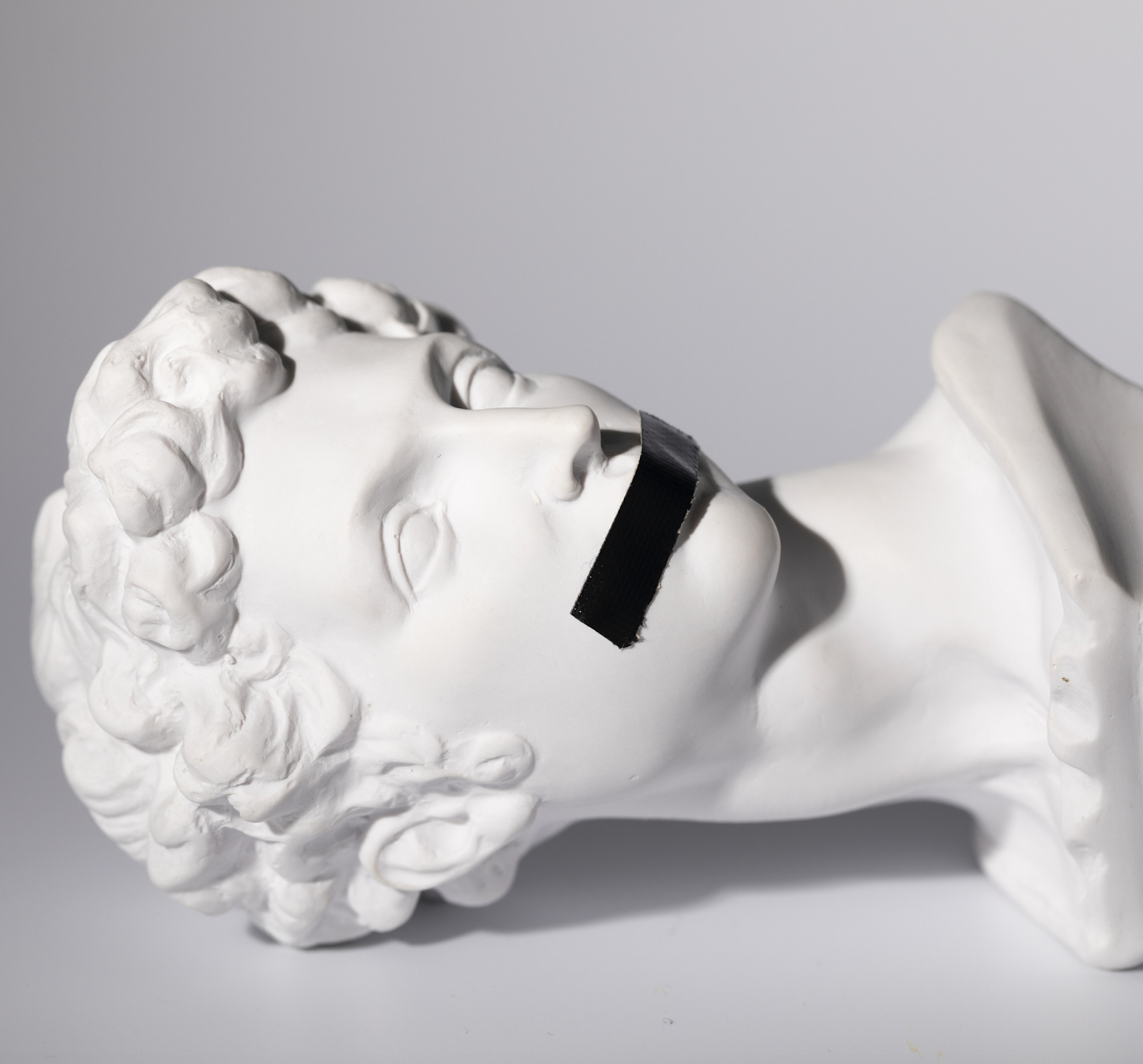 Photo of a medieval white stone sculpture of a young man's head, with black tape covering the mouth.