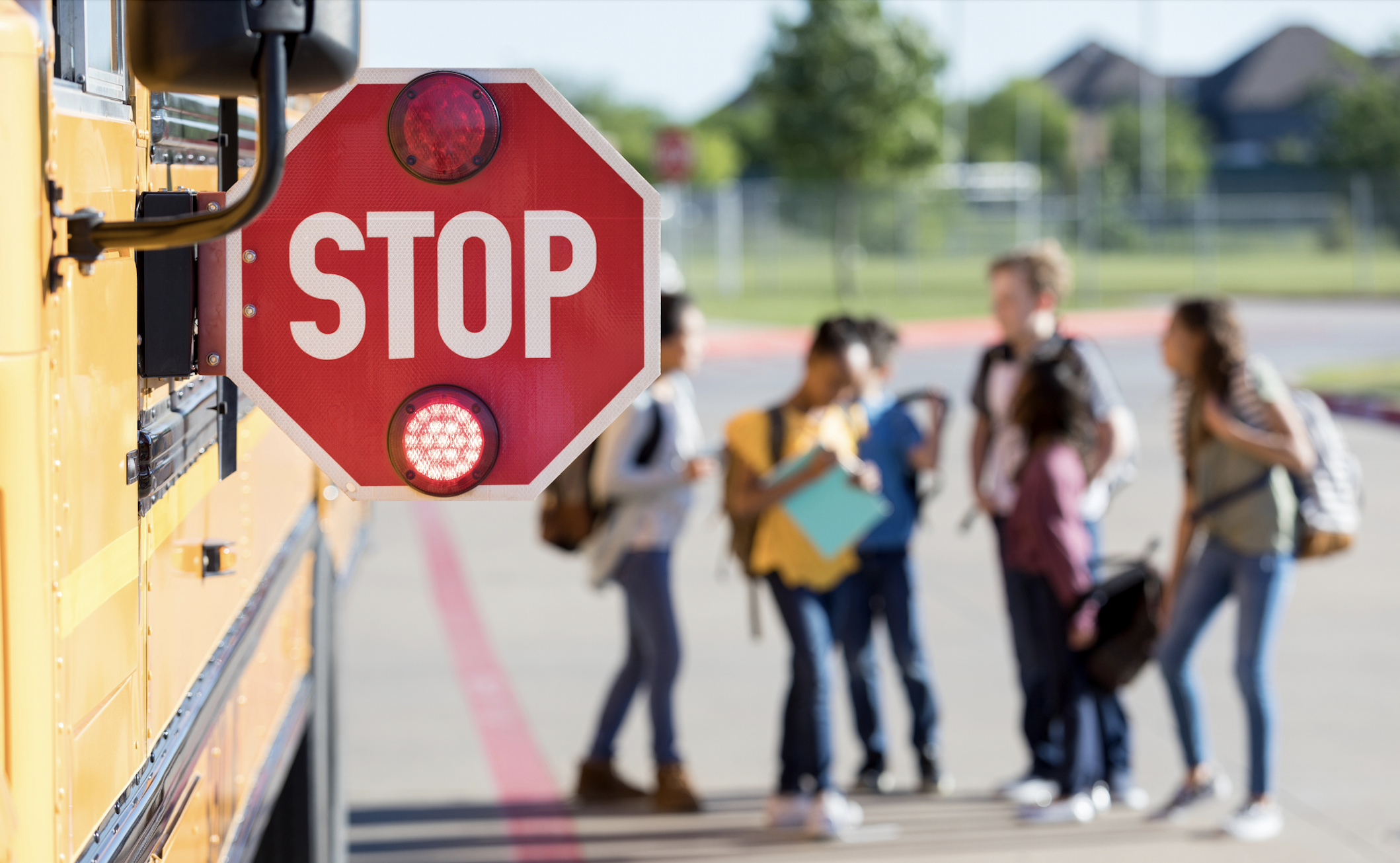 Blog featured image. Photo of a STOP sign sticking off the side of a school bus, with blurry middle-school students chatting nearby.