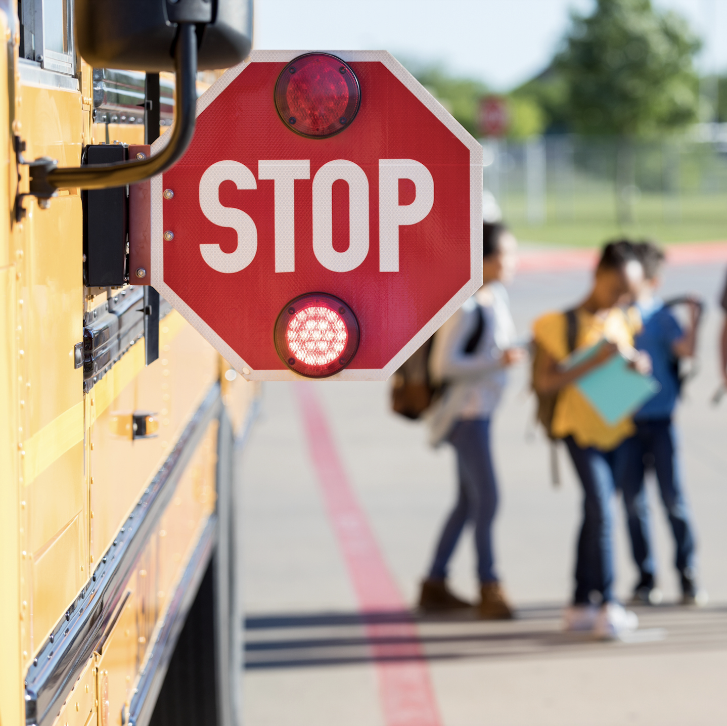 Blog featured image. Photo of a STOP sign sticking off the side of a school bus, with blurry middle-school students chatting nearby.