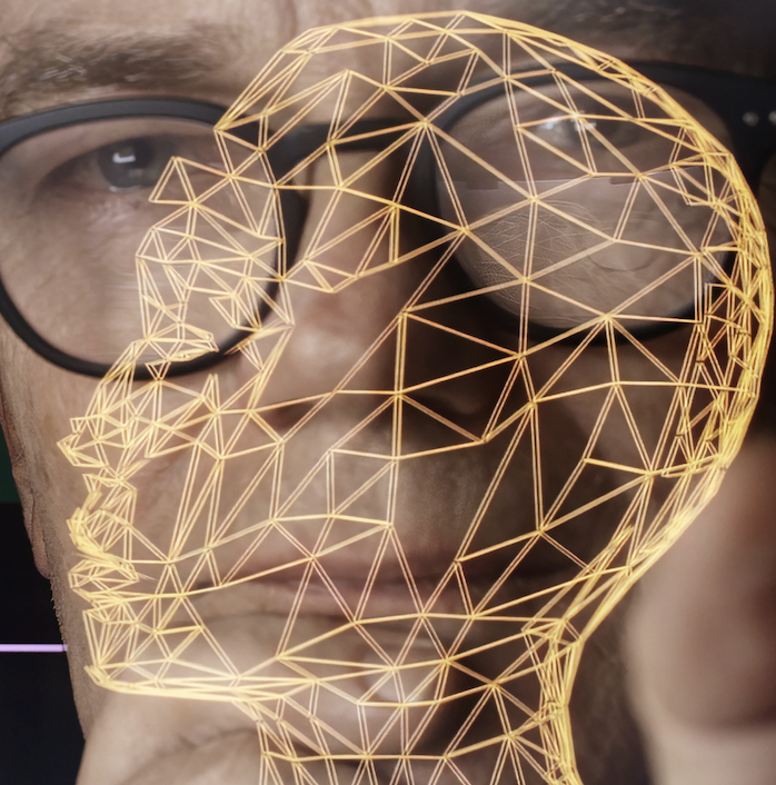 An image of a man as if seen through a touch screen as he studies graphic displays of AI avatars and coding.