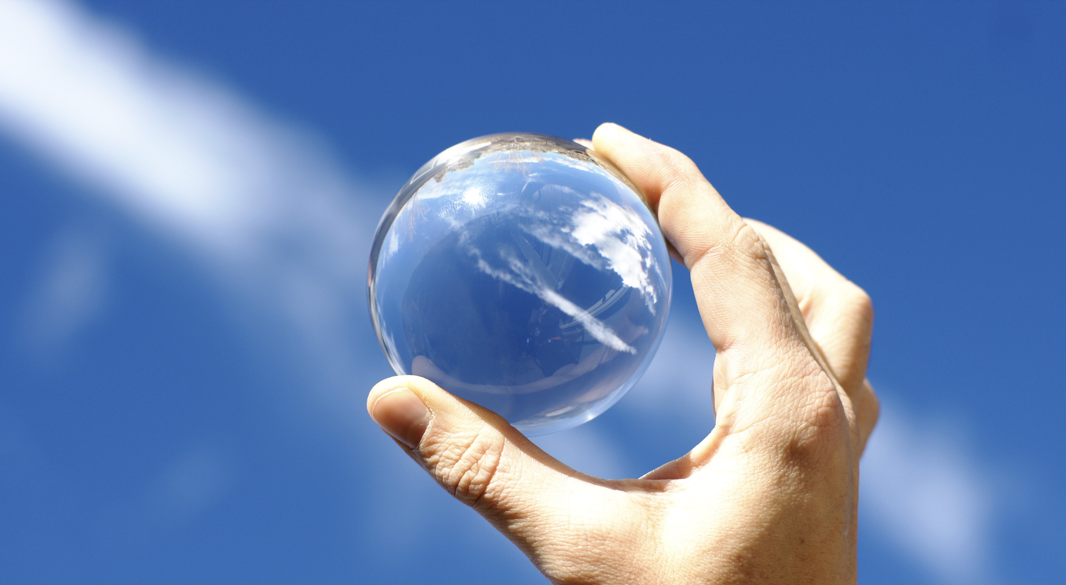 A hand holds a crystal ball aloft against a blue sky with wispy clouds.