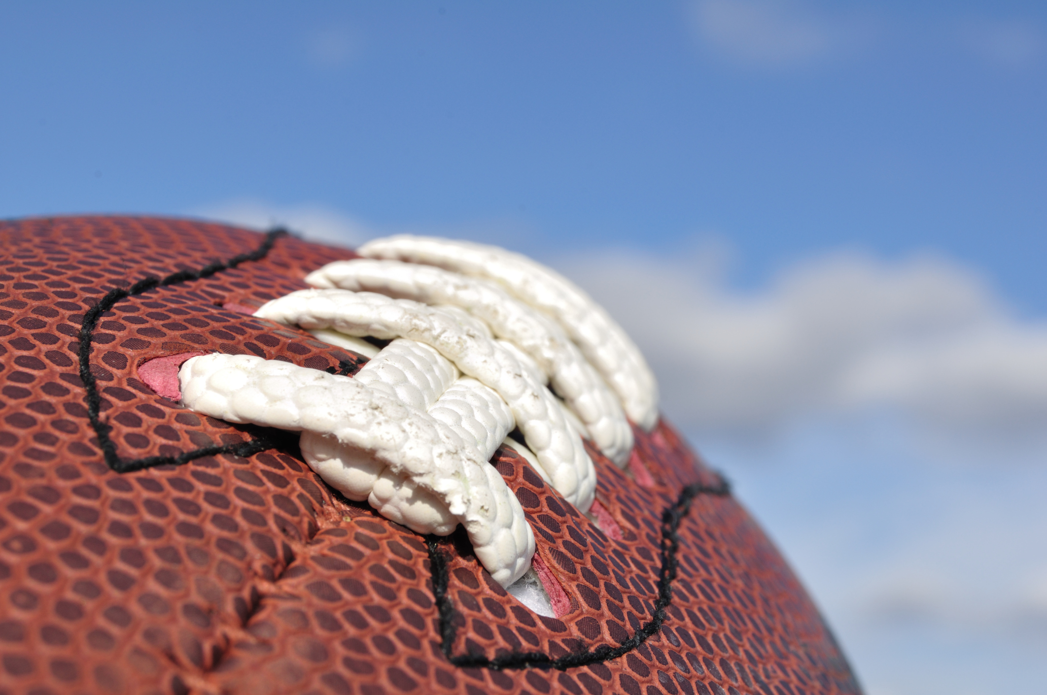 Closeup photo of a brown football with white laces and a blue sky beyond.